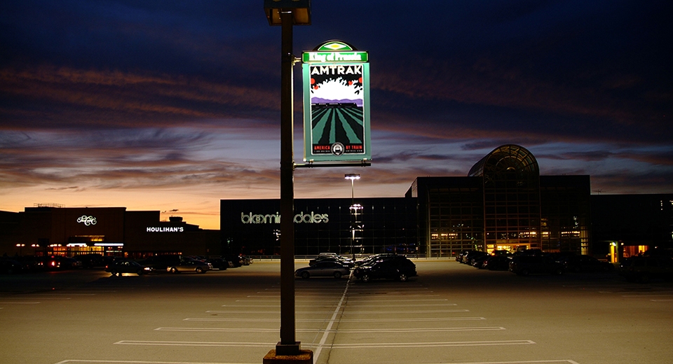 Image of a Lumen Banner displayed on a light pole in the King of Prussia parking lot. It displays an advertisement for Amtrak.
